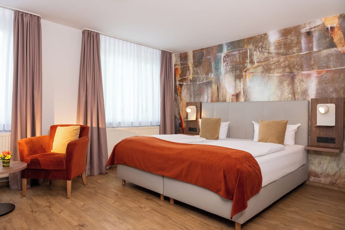 Ein Hotelzimmer mit einem großen Bett, einem Sessel, einem Tisch mit Blumen und Blick auf die Stadt durch das Fenster.