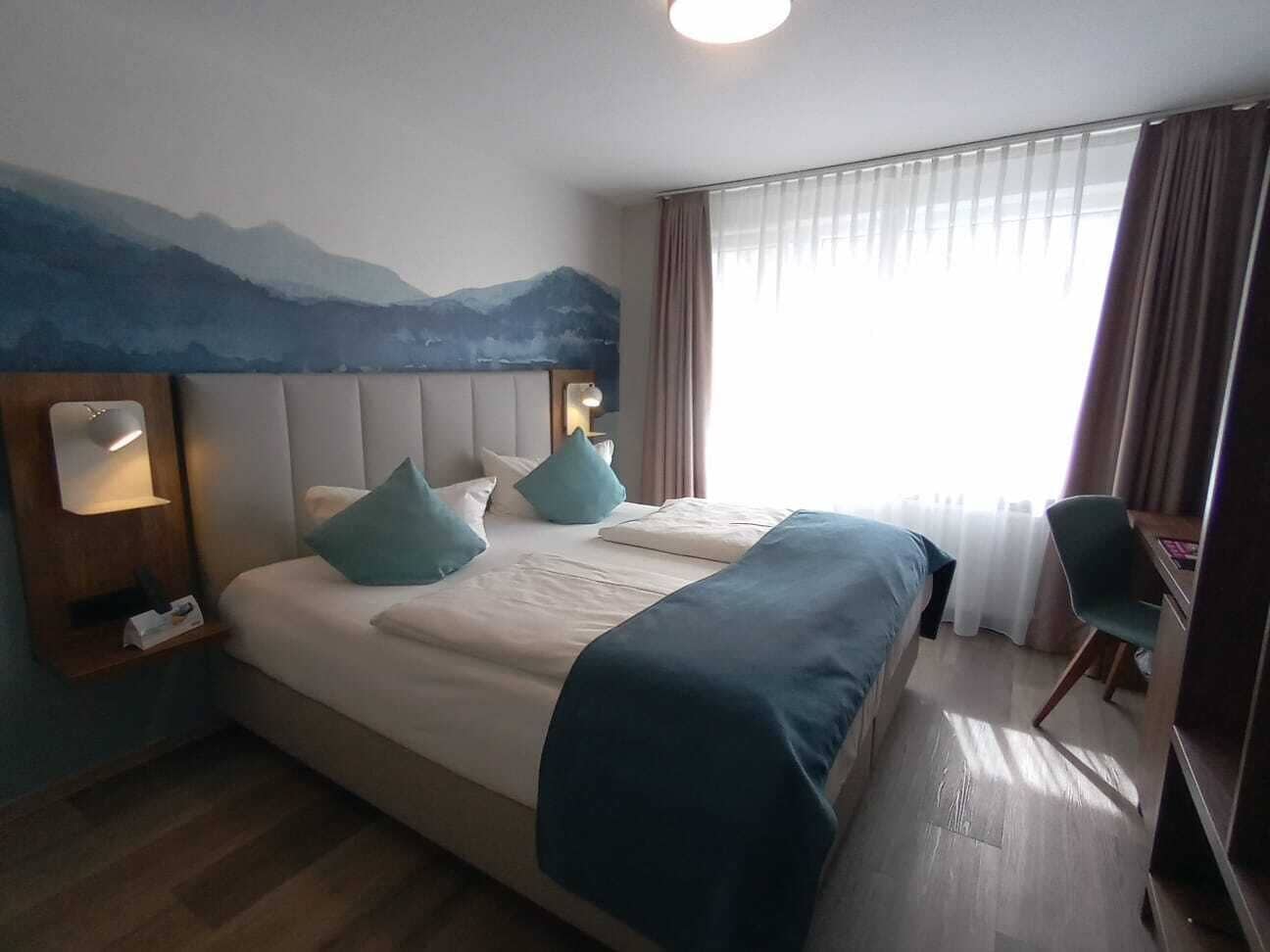 A hotel room with a bed, a desk, and a chair. There is a painting on the wall above the bed.