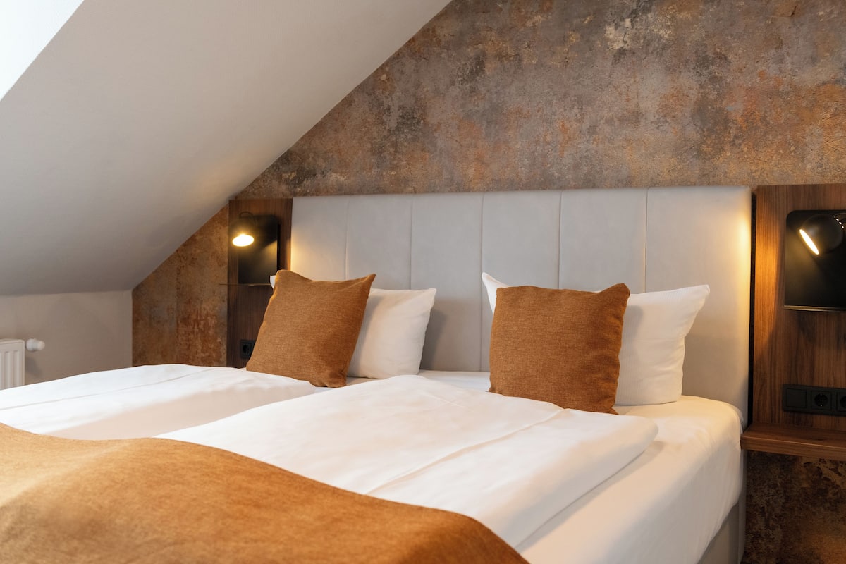 Ein Schlafzimmer mit schräg verlaufender Decke, zwei Betten mit weißer Bettwäsche und braunen Kissen, Holz-Kopfteile und Wandleuchten.