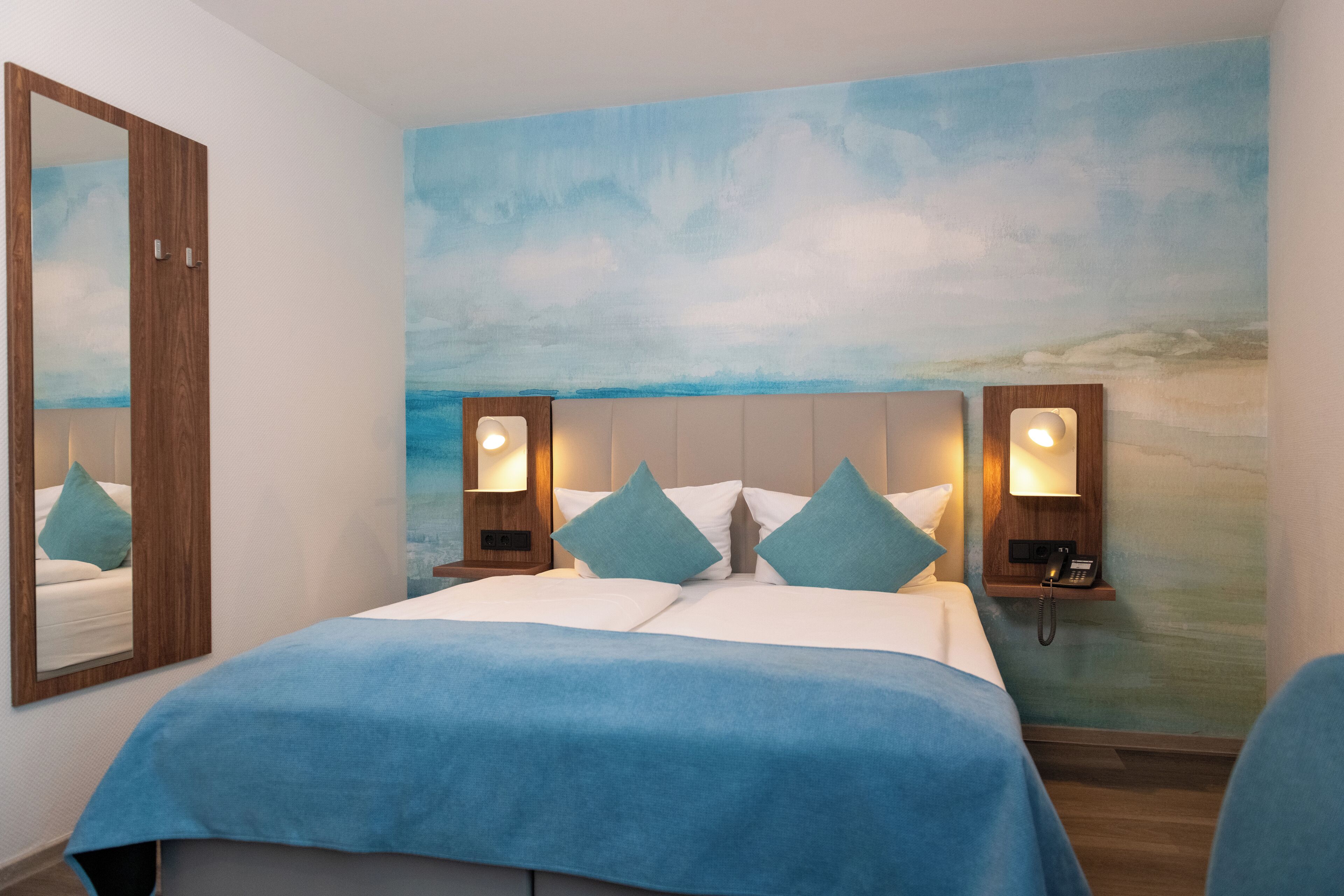 A bedroom with a bed, a nightstand, a mirror, and a wall with a painted sky and clouds.