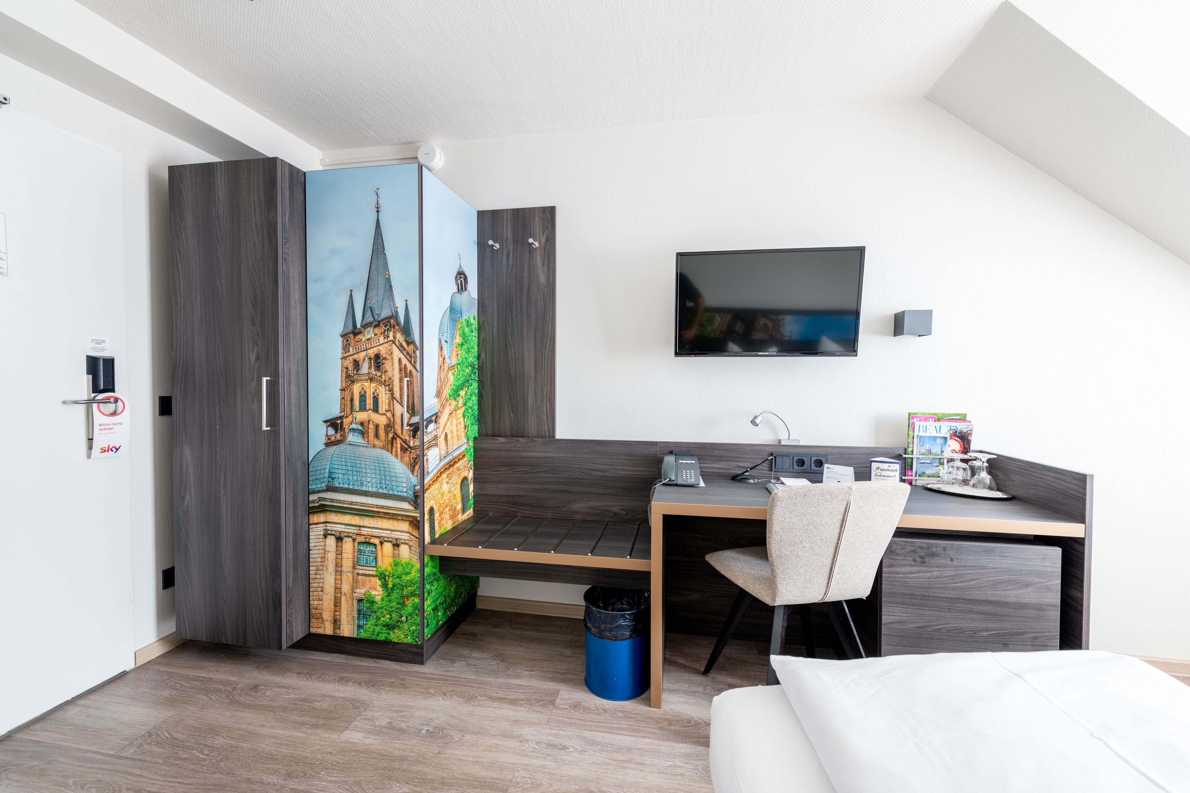 A modern hotel room with a bed, a desk with a chair, a TV mounted on the wall, and a large mural of a building.