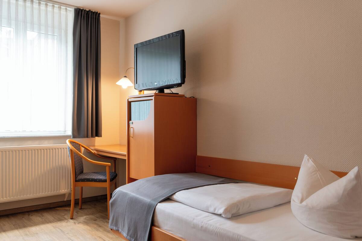 Ein Hotelzimmer mit Bett, Schreibtisch, Stuhl, Fernseher auf einem Standfuß und einem fenster mit Vorhängen.