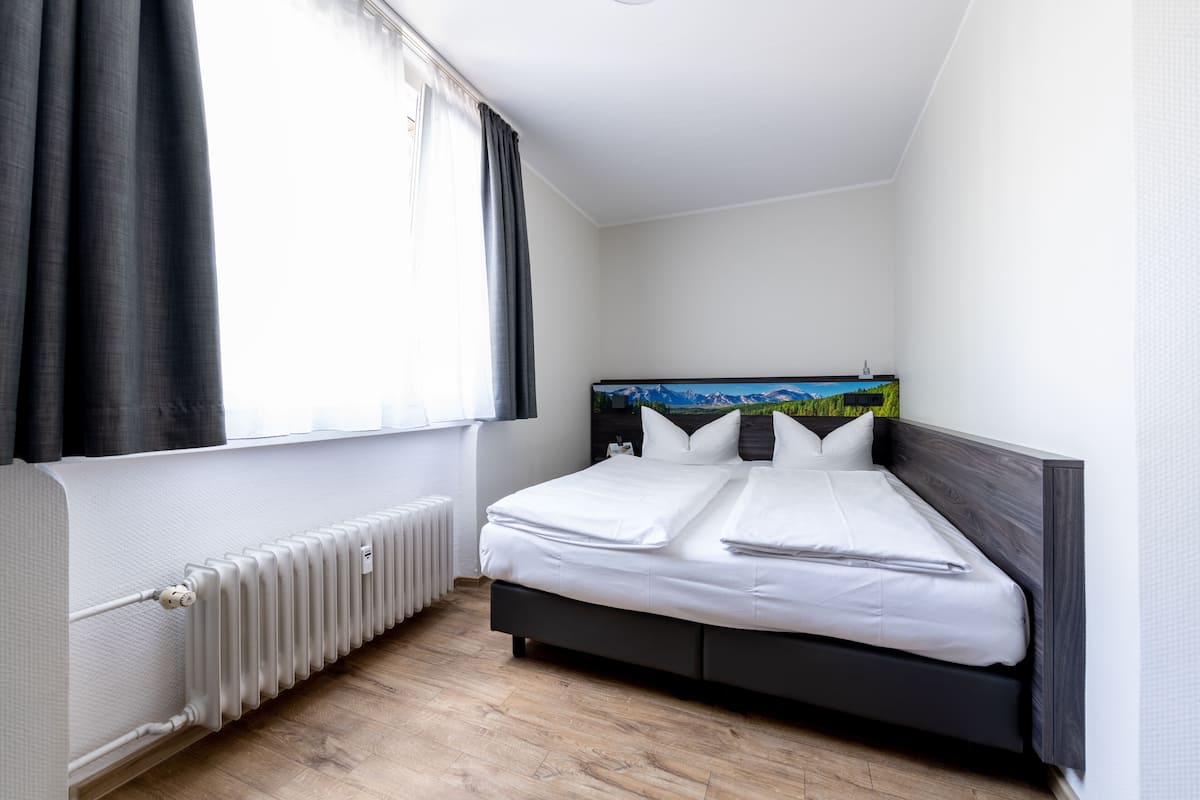 Ein ordentlich bezogenes Bett in einem Eckzimmer, mit Heizkörper, einem fenster mit Vorhängen und einem Bild an der Wand.