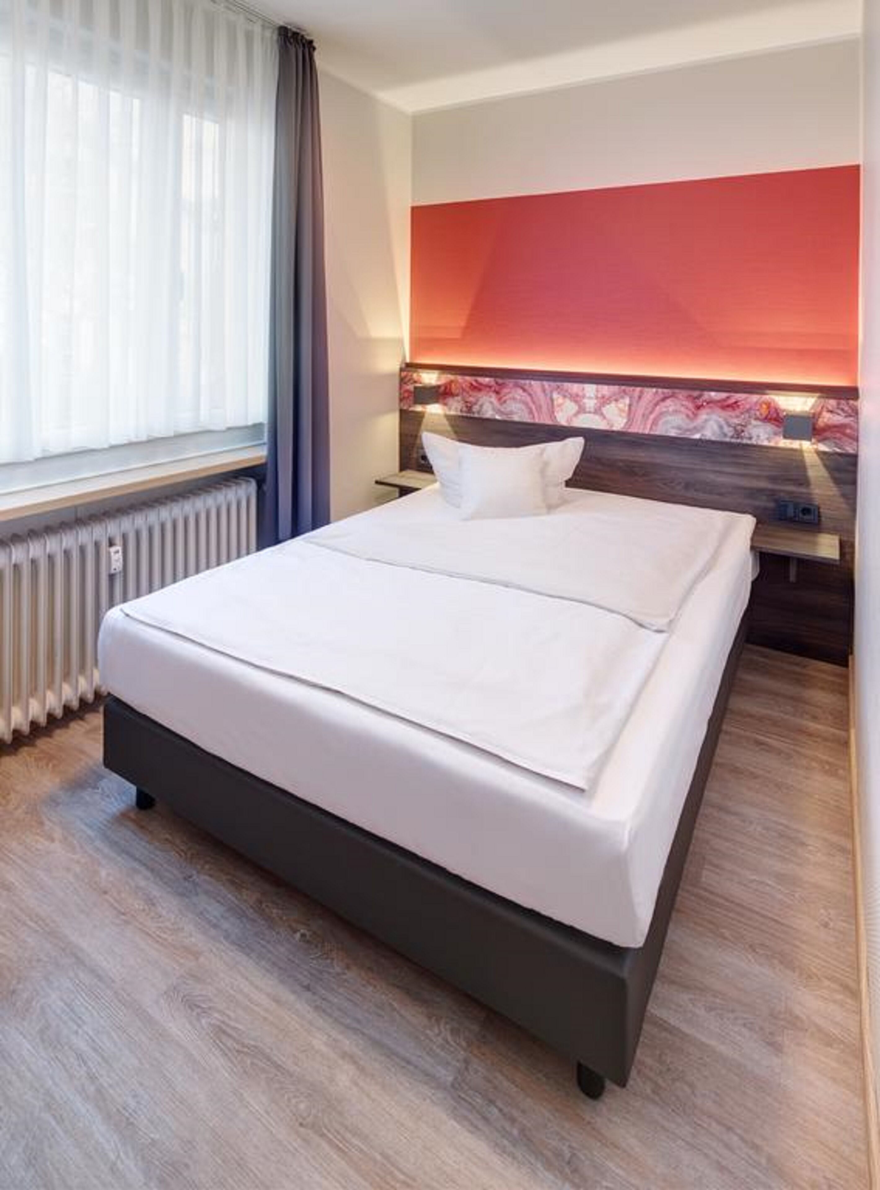 Habitación de hotel con una cama grande, un radiador, una ventana con cortinas y una pared de acento roja.