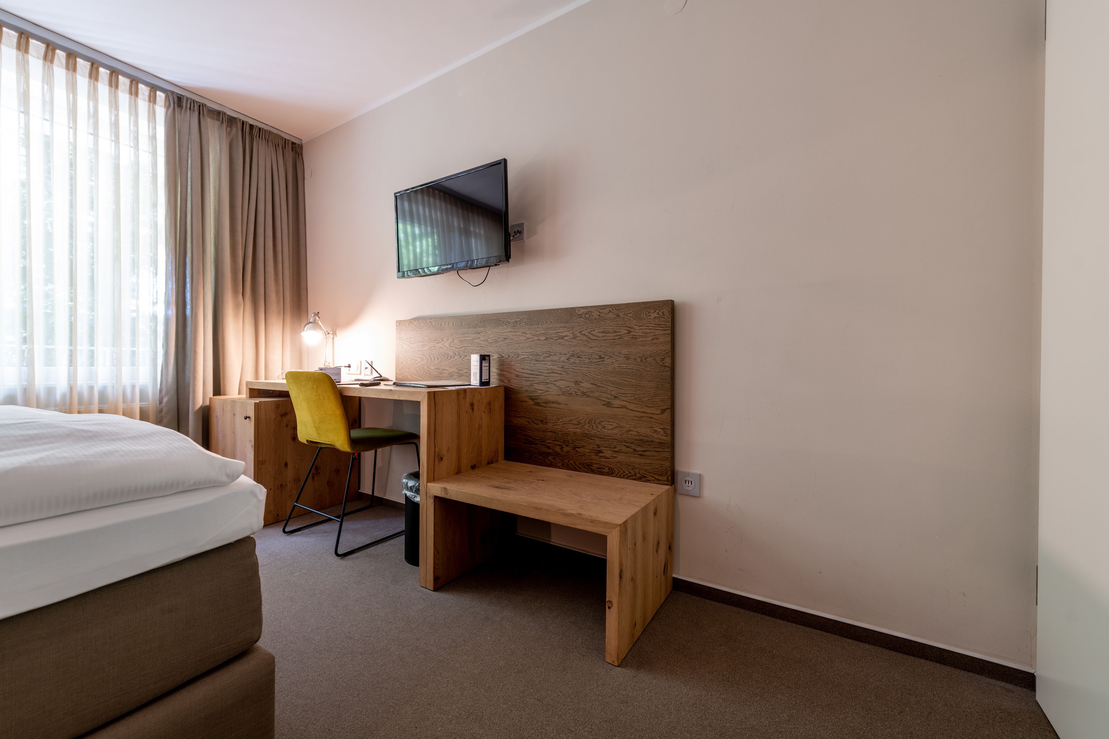 Een hotelkamer met een bed, een bureau met stoel, een aan de muur bevestigde televisie en een raam met gordijnen.