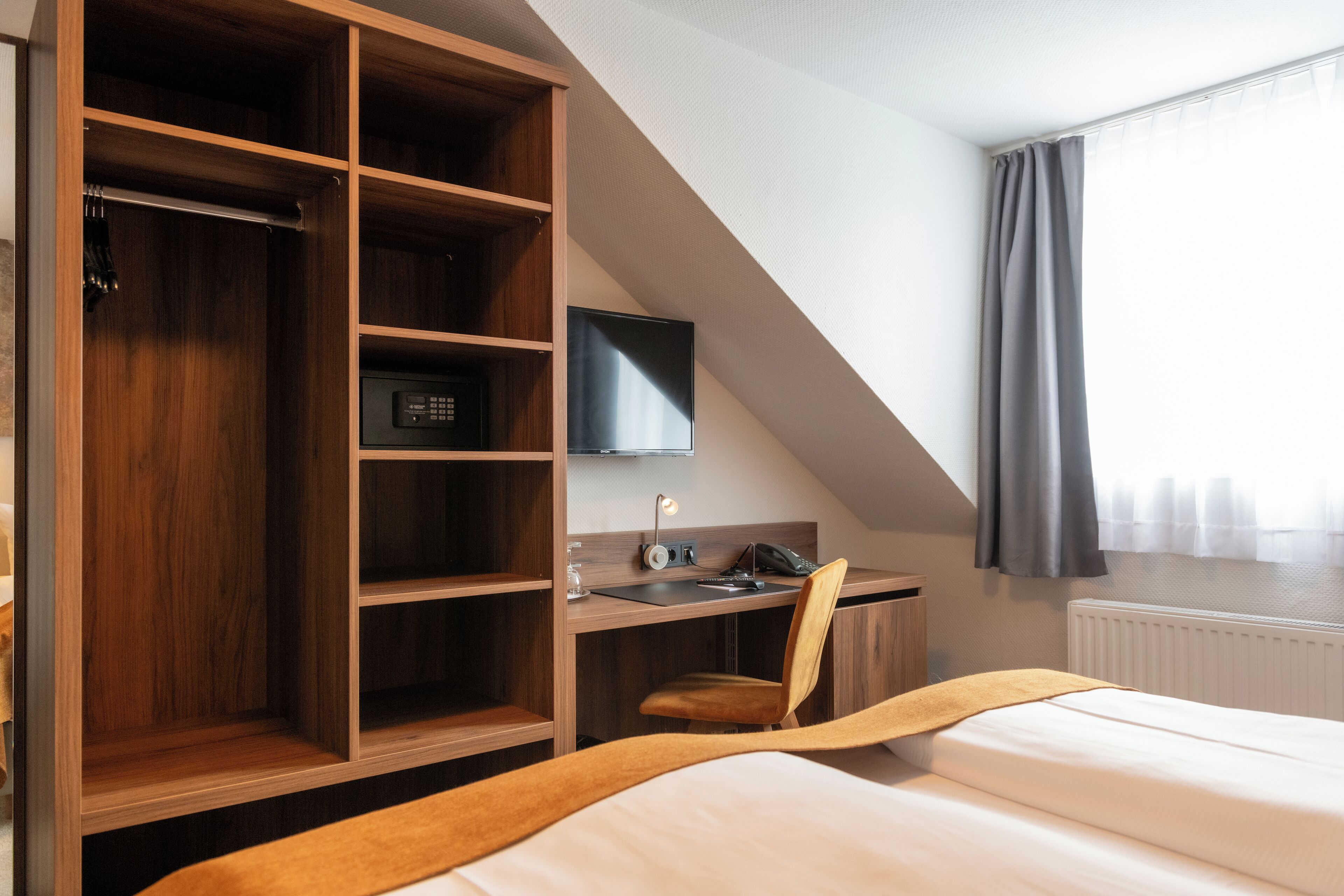 A hotel room with a wooden wardrobe, a desk with a chair, a bed, a television, and a window with curtains.