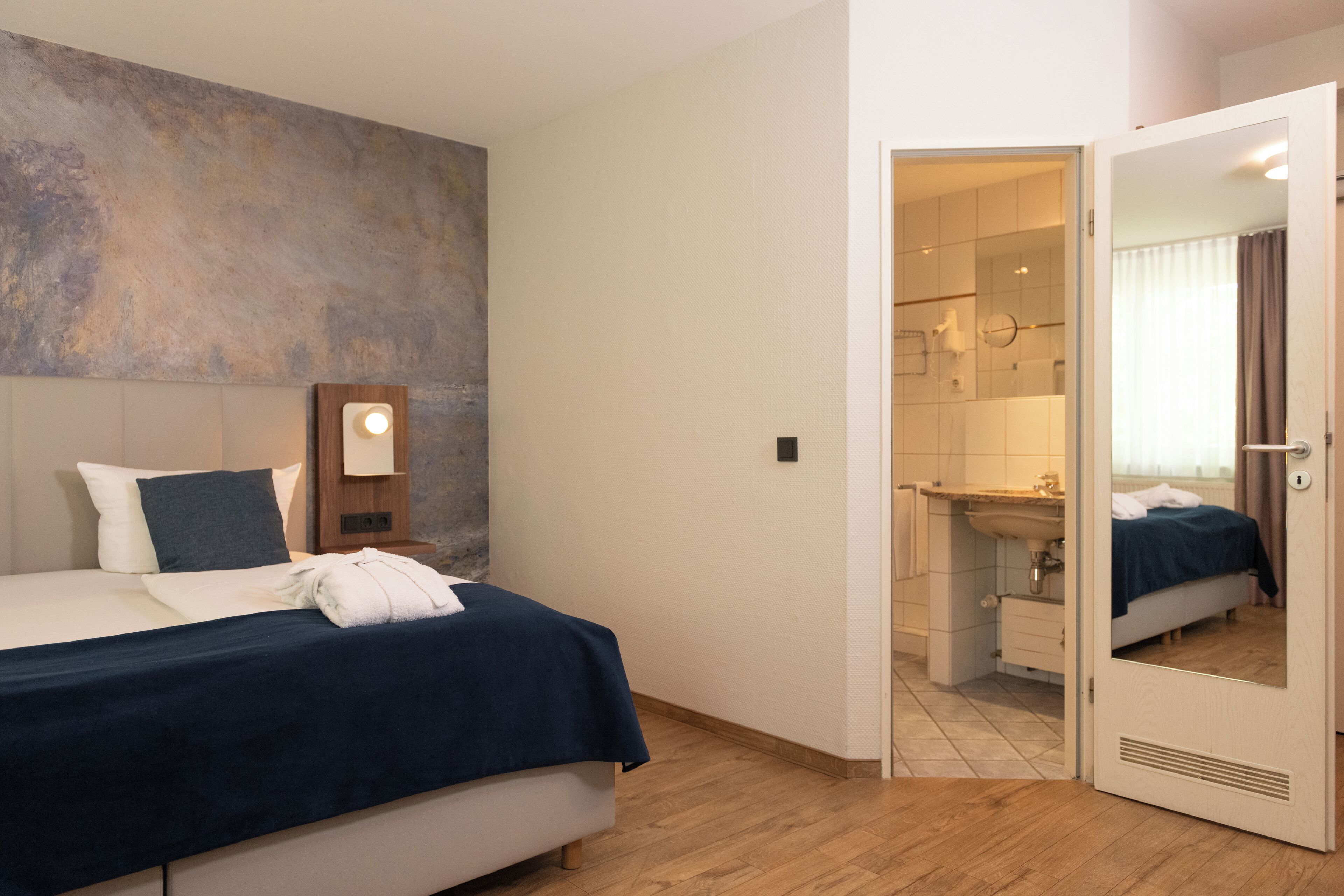 Een hotelkamer met een bed, nachtkastje, spiegel en een zichtbare badkamer door een geopende deur.