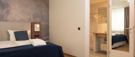Habitación de hotel con cama, mesita de noche, espejo y baño visible a través de una puerta abierta.