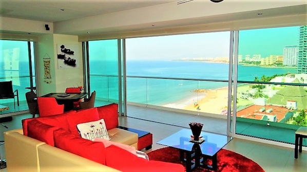 Un salon moderne avec un canapé rouge, une petite table et une grande fenêtre donnant sur la plage et le paysage urbain.