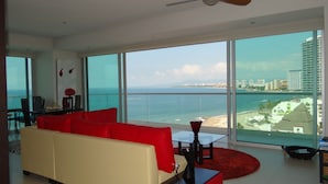 A modern living room with a red sofa, a small table, and a red rug. Large windows offer a view of the ocean and a cityscape.