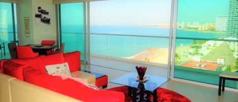 A modern living room with a red sofa, a glass coffee table, and a view of the beach and cityscape through large windows.
