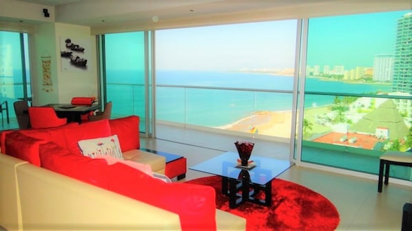 A modern living room with a red sofa, a glass coffee table, and a view of the beach and cityscape through large windows.