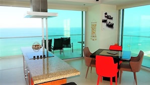 A modern kitchen with a large island, dining area with red chairs, and a view of the ocean.