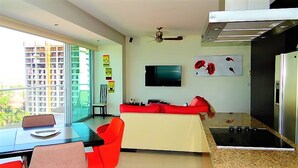 A modern kitchen with a dining area, a TV mounted on the wall, and a balcony with a view of buildings.