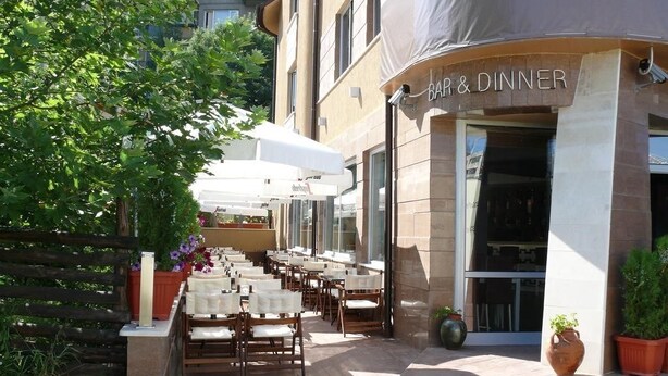 Un espace extérieur aménagé pour les repas, avec des tables et des chaises, un bâtiment portant une enseigne « BAR ET RESTAURANT », et des plantes en pot.