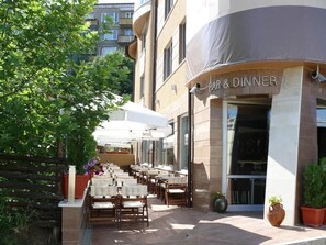 An outdoor dining area with tables and chairs, a building with a 'BAR & DINNER' sign, and potted plants.