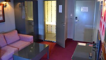 Camera d'albergo con un divano rosa, un tavolo con piano in vetro, un armadio, una zona doccia e una porta con un cartello.
