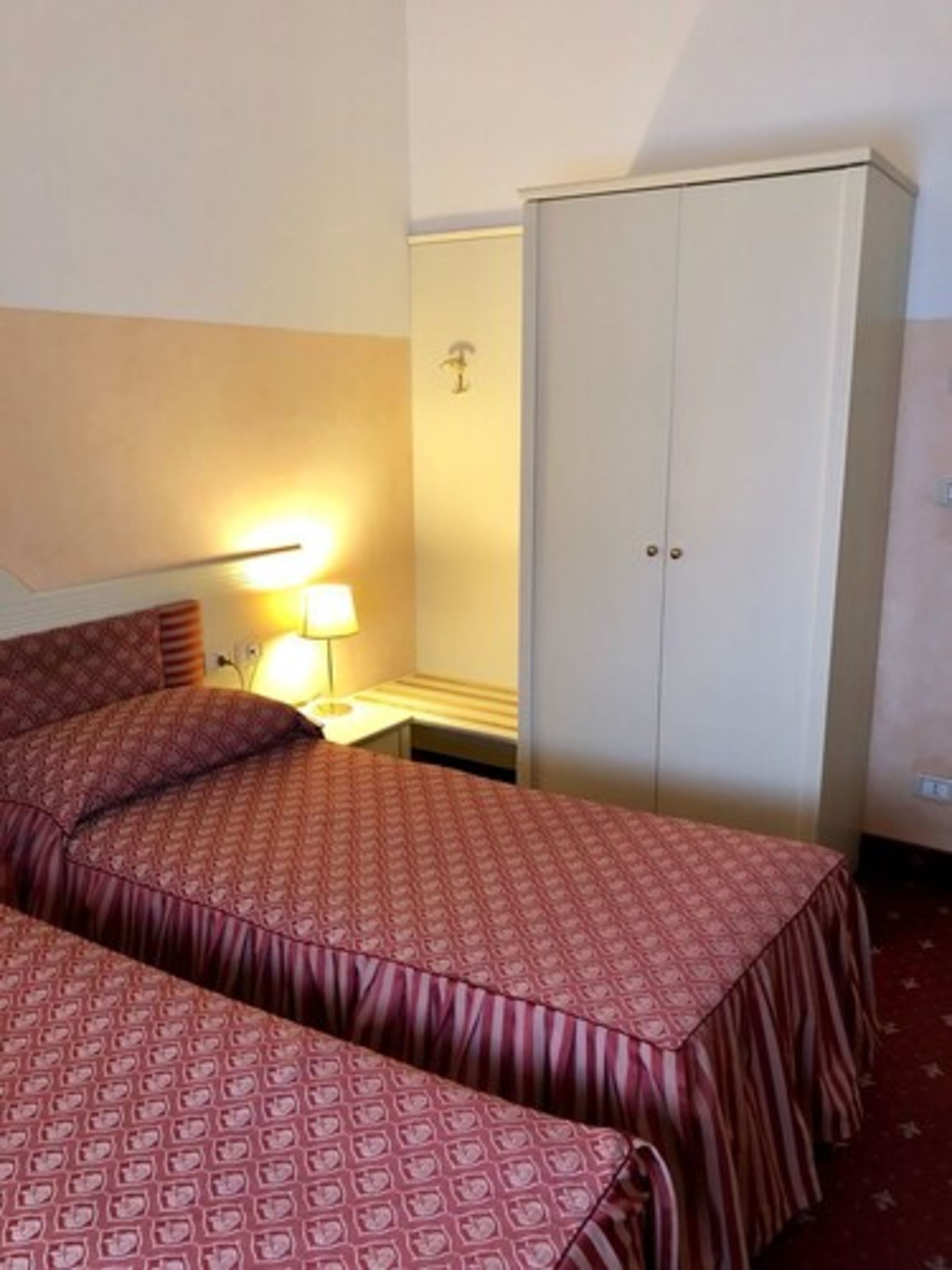 A hotel room with two beds, a bedside table with a lamp, and a wardrobe.