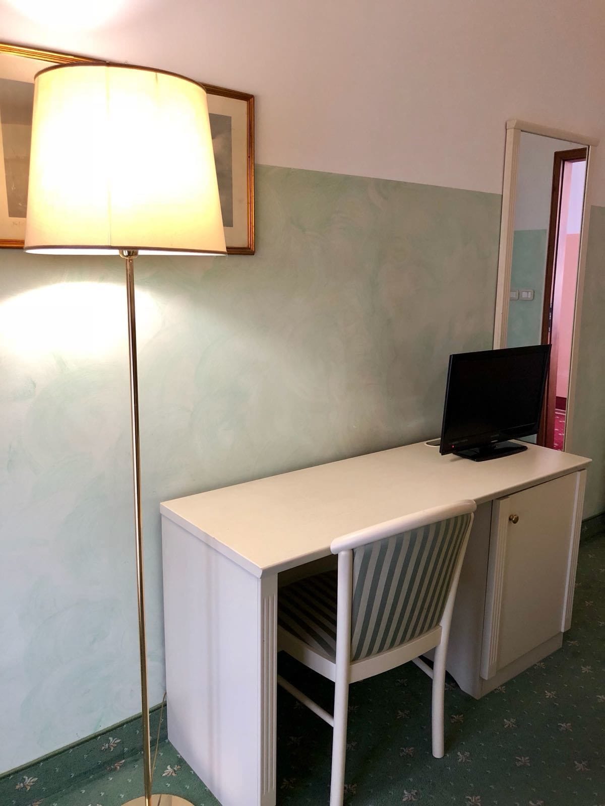 Una habitación pequeña y privada con escritorio, una silla, televisor y lámpara de pie.