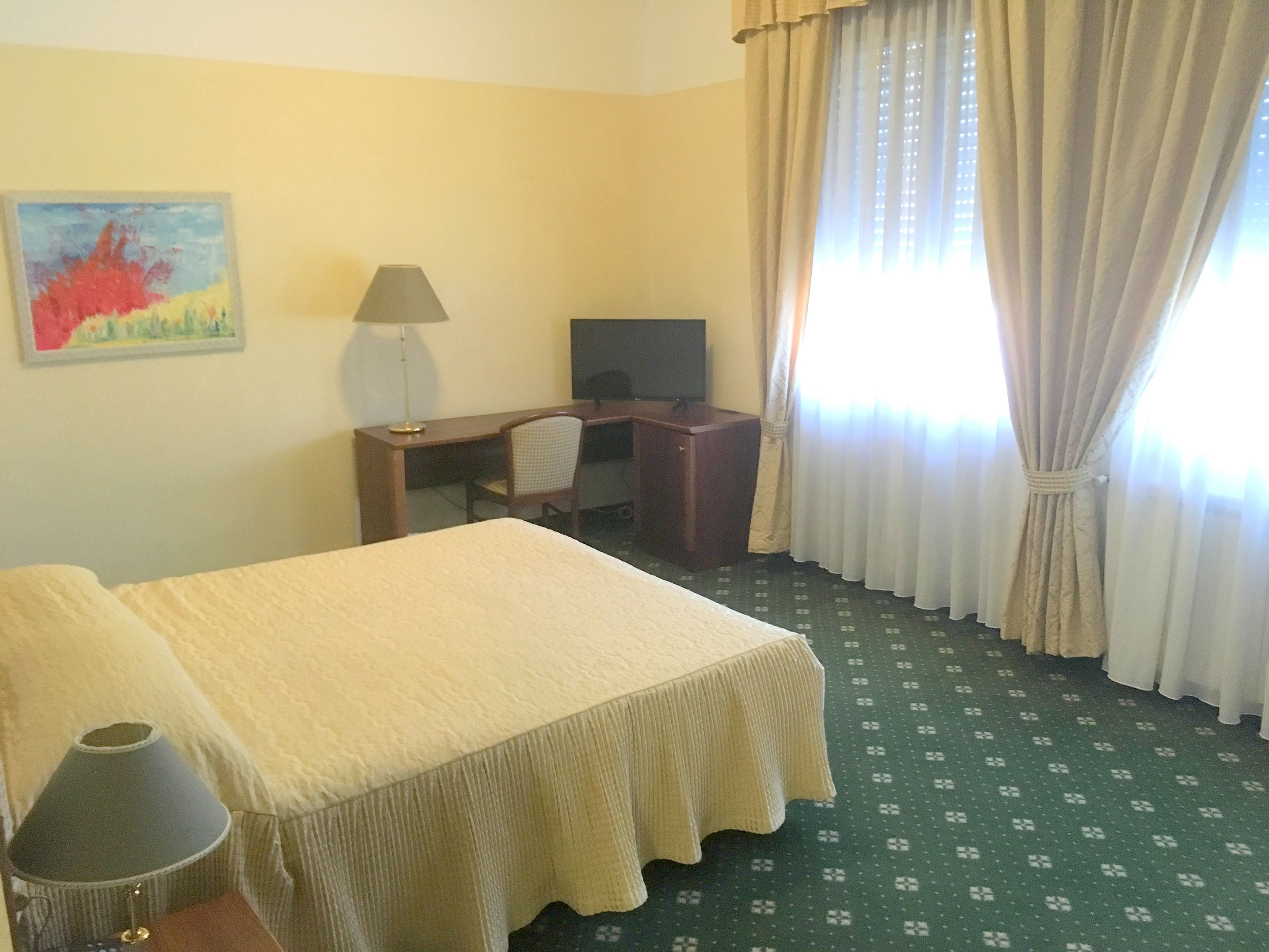 Una habitación de hotel con cama, escritorio, silla, lámpara, televisor y cortinas.