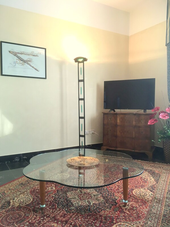 Una sala de estar con una mesa de vidrio, un televisor de pantalla plana, un armario de madera, un cuadro enmarcado en la pared y una lámpara de pie alta.
