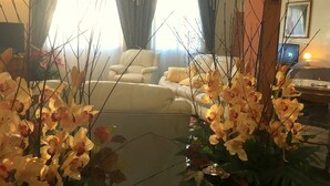 A living room with a sofa, a coffee table, and a lamp. There is a window with curtains and a plant in the foreground. - Hotel Internazionale (Gorizia)