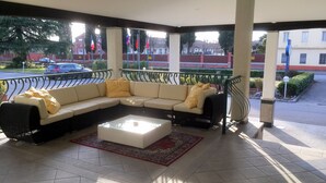 A covered outdoor seating area with a sectional sofa and a coffee table, featuring a view of the street and buildings in the background. - Hotel Internazionale (Gorizia)
