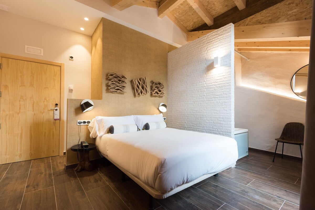 A modern bedroom with a large bed, wooden ceiling beams, a white brick accent wall, and a small bedside table with a lamp.
