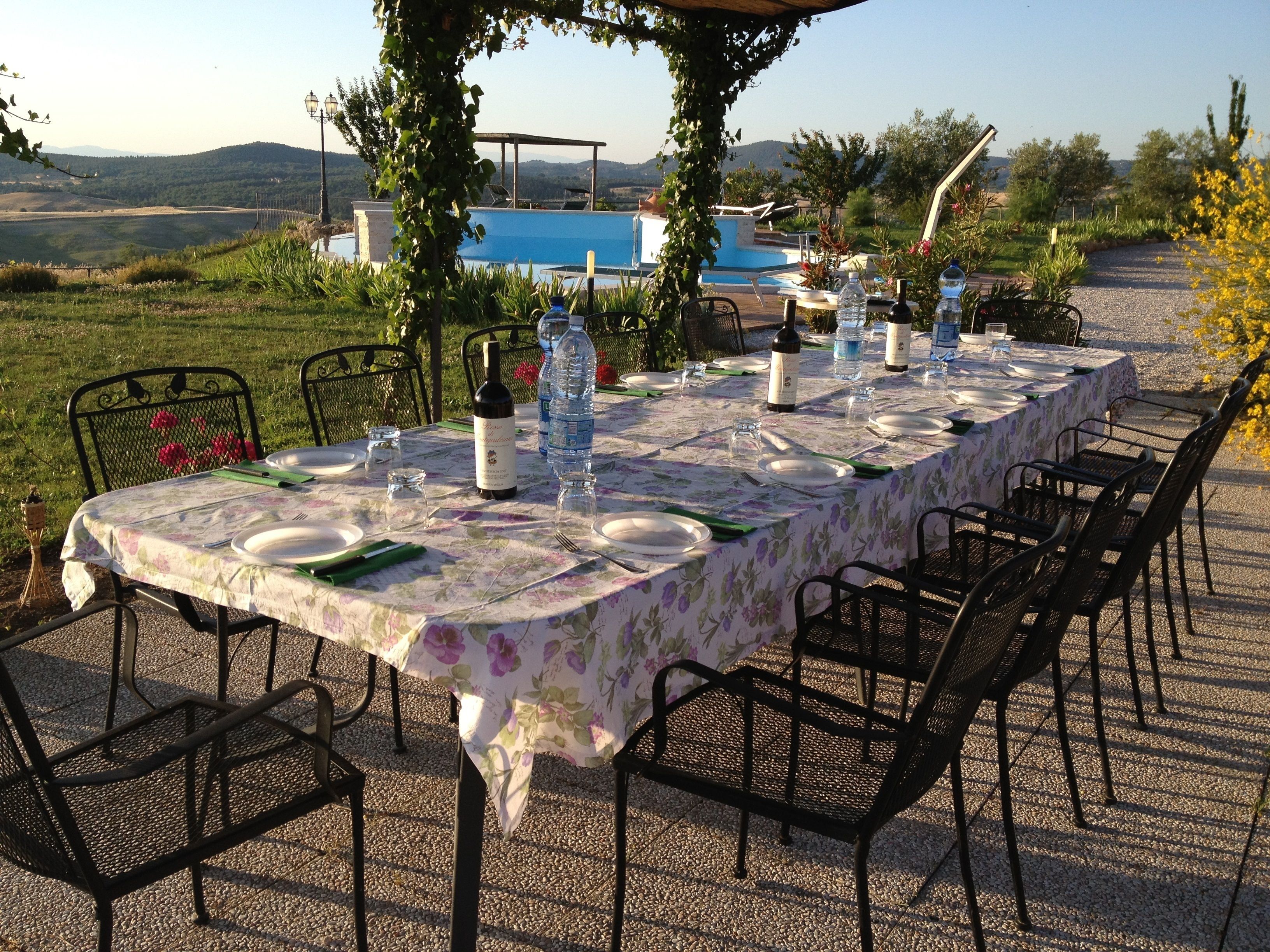 Un área de comedor al aire libre con una mesa dispuesta para seis personas, rodeada de sillas, con una piscina y colinas al fondo.