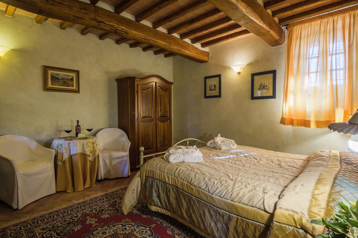 A bedroom with a large bed, wooden ceiling beams, a wooden wardrobe, two chairs, a small table with a tablecloth, and a red patterned rug.