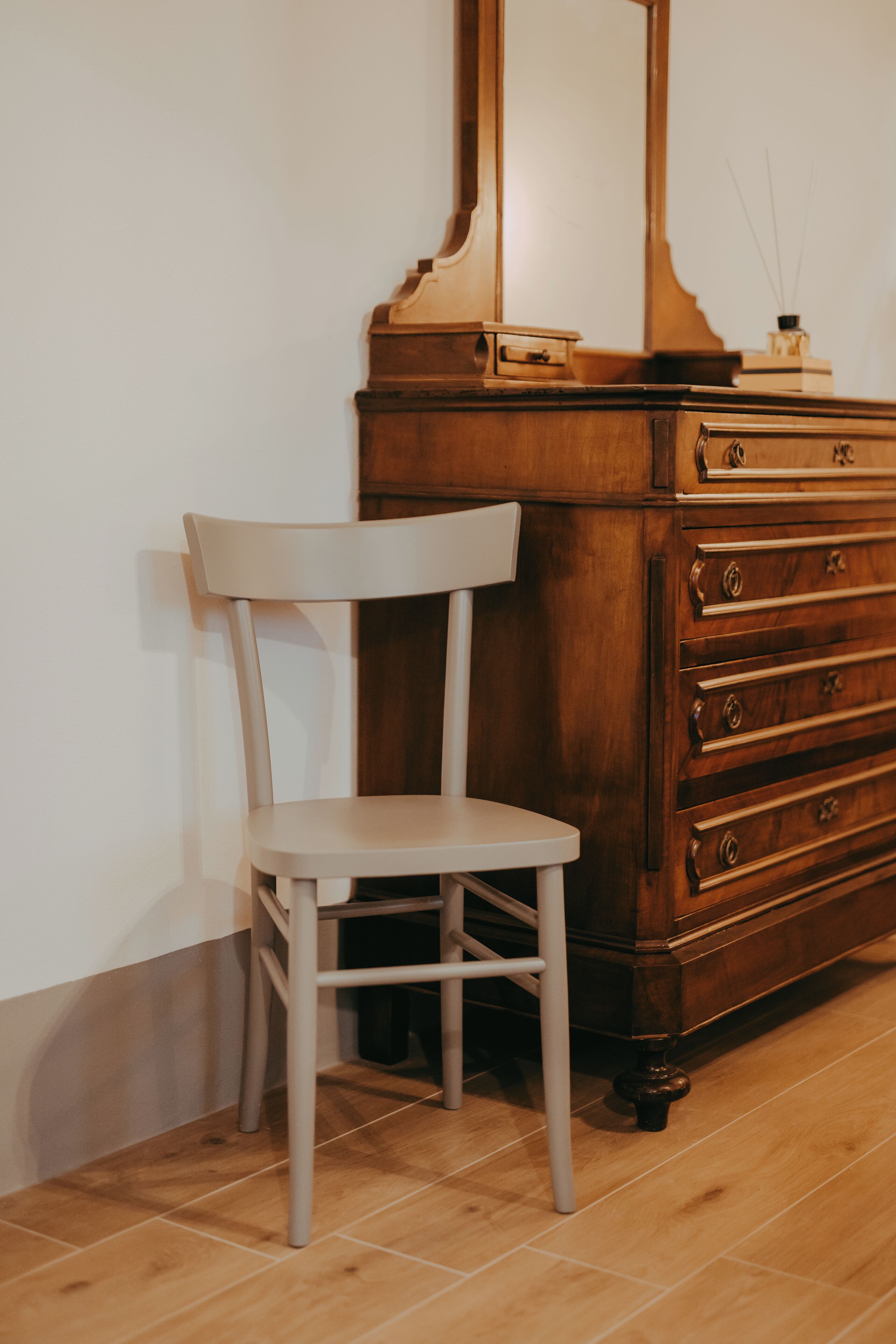 Un aparador de madera con cajones y un espejo, una silla blanca y un suelo de baldosas.