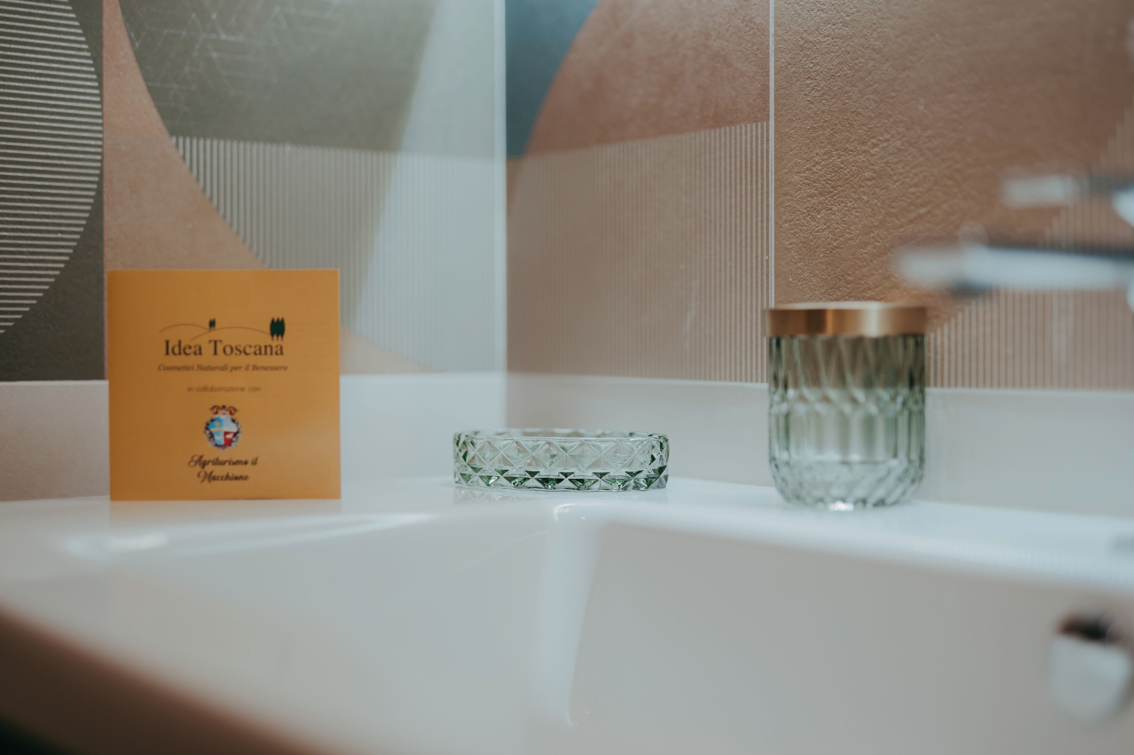 A bathroom with a white sink, a glass candle holder, and a card labeled "Idea Toscana."