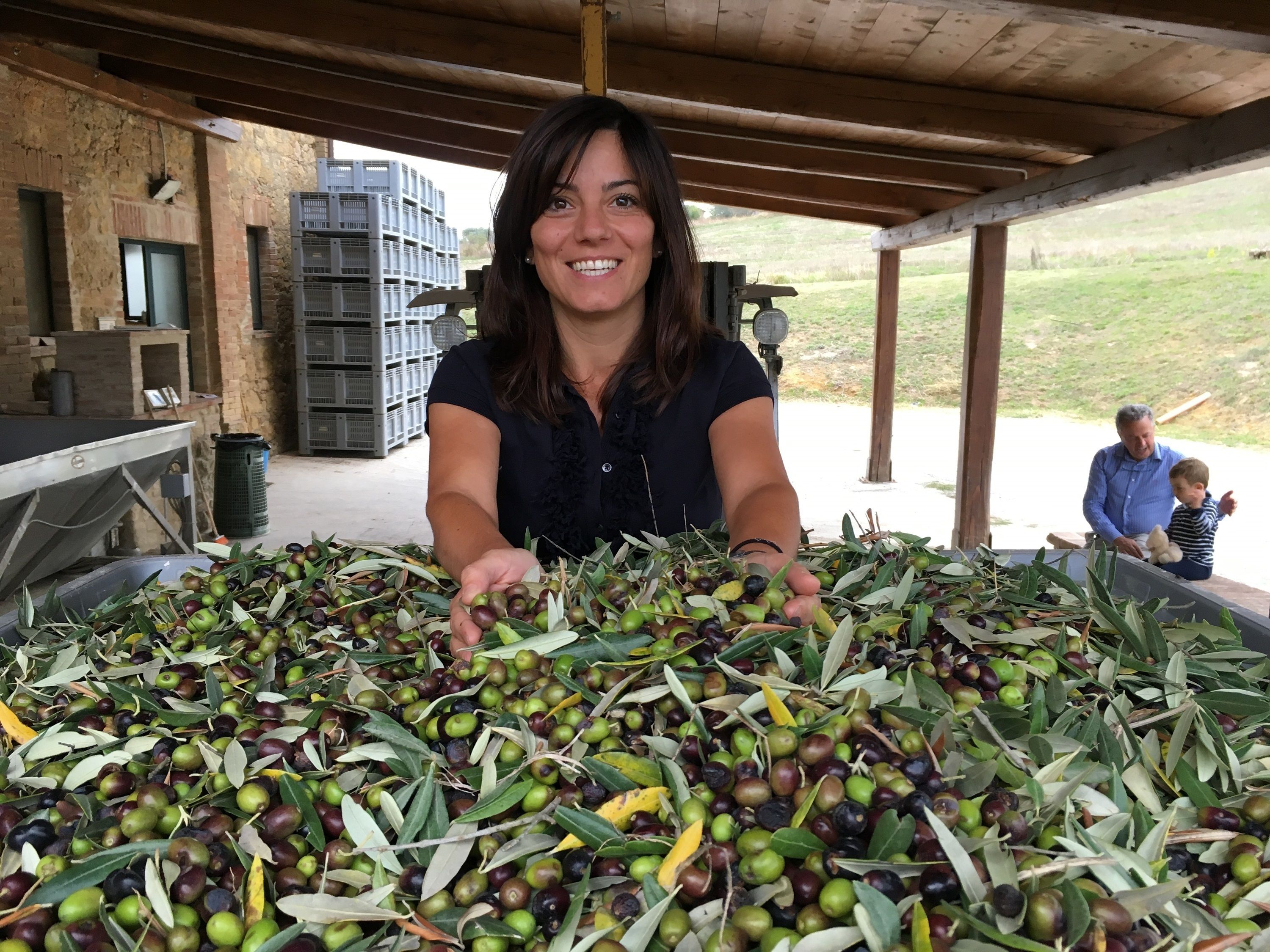Una donna maneggia olive e rami d'ulivo in un'area esterna coperta, con un muro in mattoni e un uomo sullo sfondo.