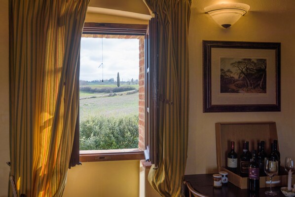 A room with a window, curtains, a framed picture, and a table with wine bottles and glasses. - Agriturismo Il Macchione (Pienza)