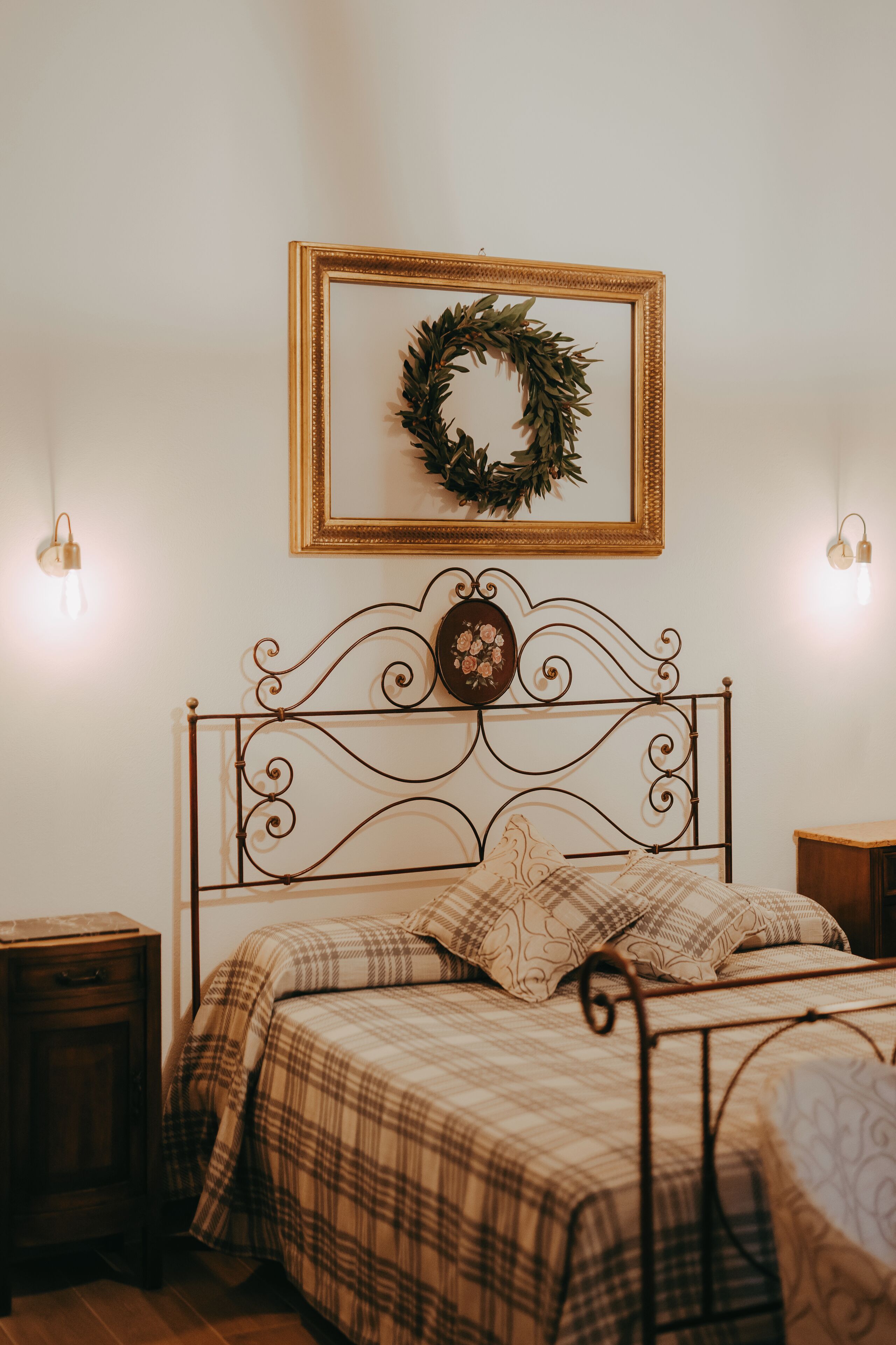 Ein Schlafzimmer mit einem Metallbettgestell, einer Karo-Bettdecke, einem gerahmten Kranz an der Wand und zwei Nachttischlampen.