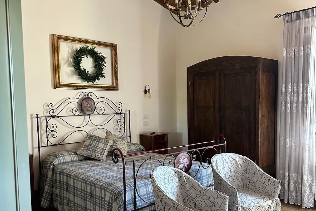 A bedroom with a bed, two chairs, a wardrobe, a chandelier, a framed wreath, and a wall-mounted light.