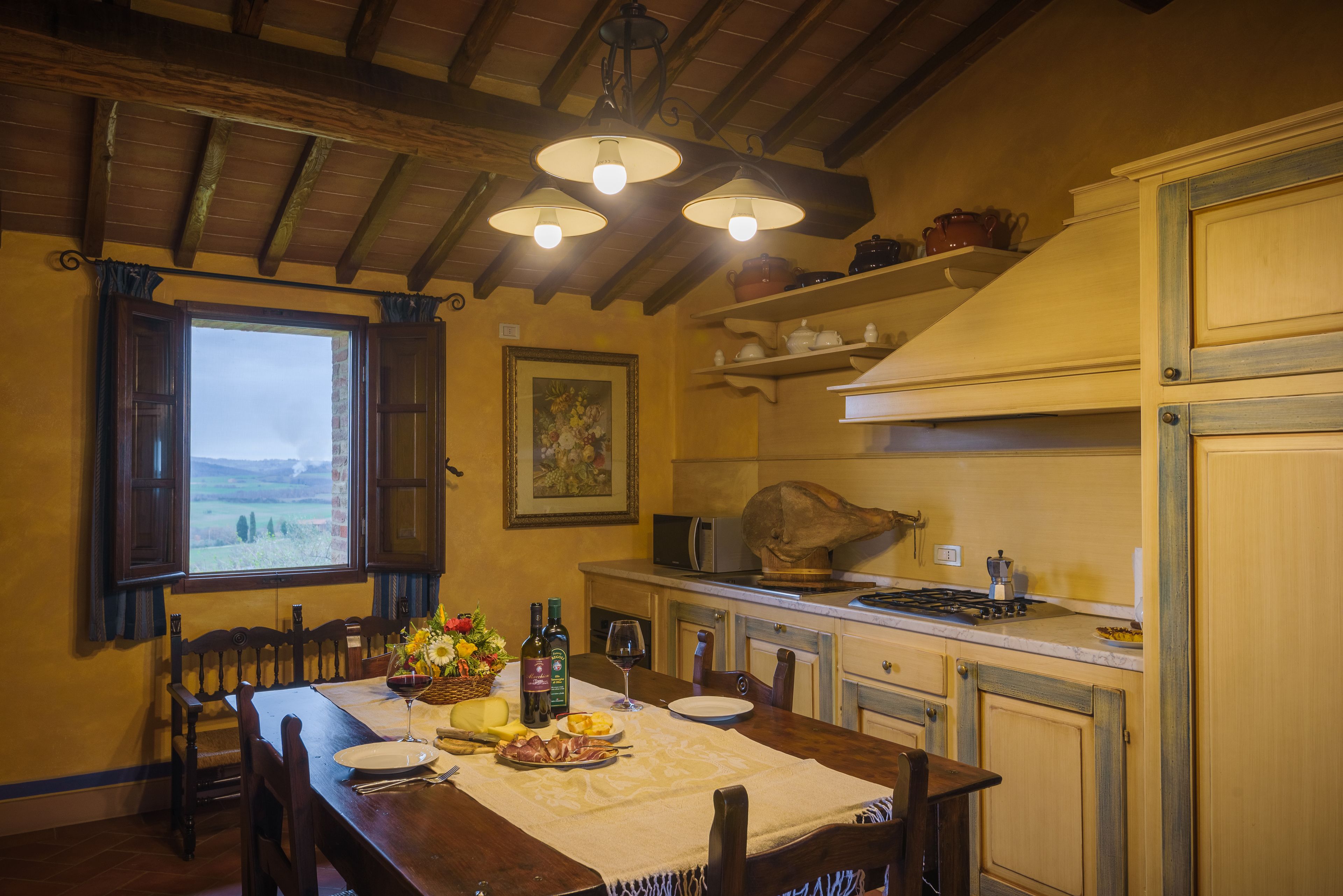 Un comedor con muebles de madera, una mesa dispuesta con platos, una botella de vino y una cesta de flores. Hay una ventana con contraventanas abiertas, un cuadro en la pared y un microondas en la encimera.