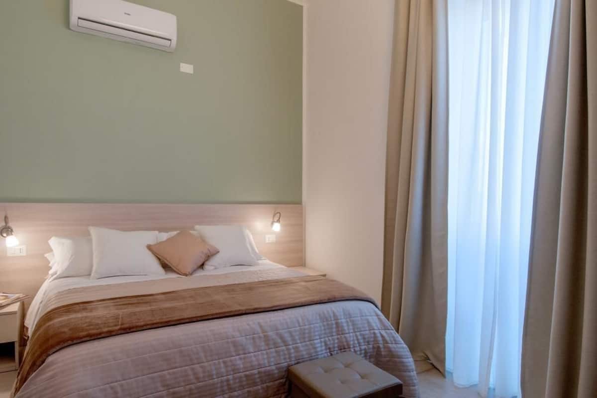 Una cama bien tendida con cabecera, una lámpara de noche y un aire acondicionado de pared.
