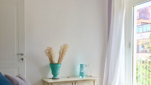 A bedroom with a blue sofa, a white door, a small white dresser, and a window with white curtains.