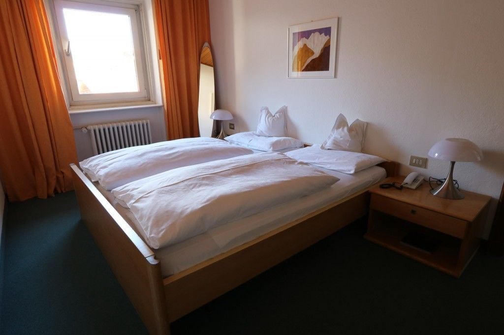 Un lit double avec du linge de lit blanc, deux tables de chevet avec des lampes, un tableau encadré au mur et une fenêtre avec des rideaux.