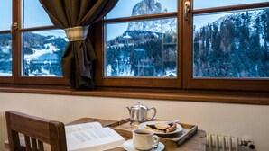 Una mesa de madera con un libro, una tetera, una taza de café y un plato de comida junto a una ventana que ofrece vistas de montañas nevadas.