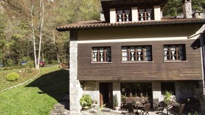 Una casa a due piani con fondamenta in pietra, rivestimento in legno e tetto in tegole. C'è un patio con mobili da esterno e un vialetto in pietra che conduce all'ingresso.