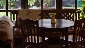 Zona pranzo con tavolo rotondo, sedie e un vaso di fiori. Grandi finestre offrono vista su un giardino con diverse piante e una recinzione in legno.