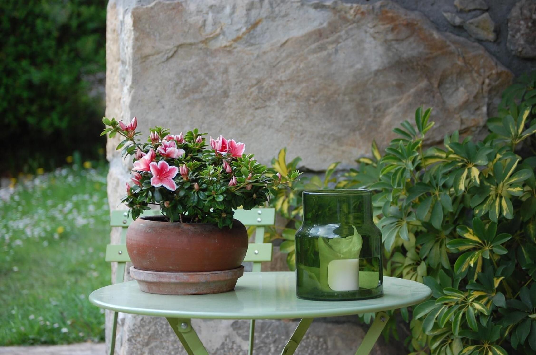 A green metal table with a terracotta pot containing pink flowers and a green glass candle holder.