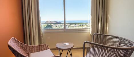 Una habitación con dos sillones de mimbre, una pequeña mesa redonda y un amplio ventanal con vistas al mar y al paisaje distante.
