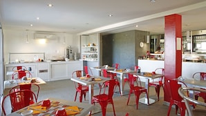 Un espace repas moderne avec des chaises rouges et des tables blanches, une cuisine en arrière-plan et un mur d’accent rouge.
