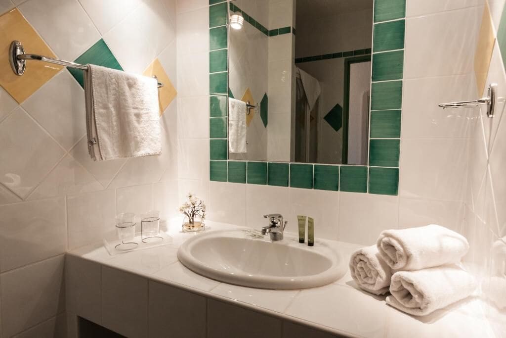 Un baño con un lavamanos blanco, azulejos verde y blanco, un perchero y un espejo.