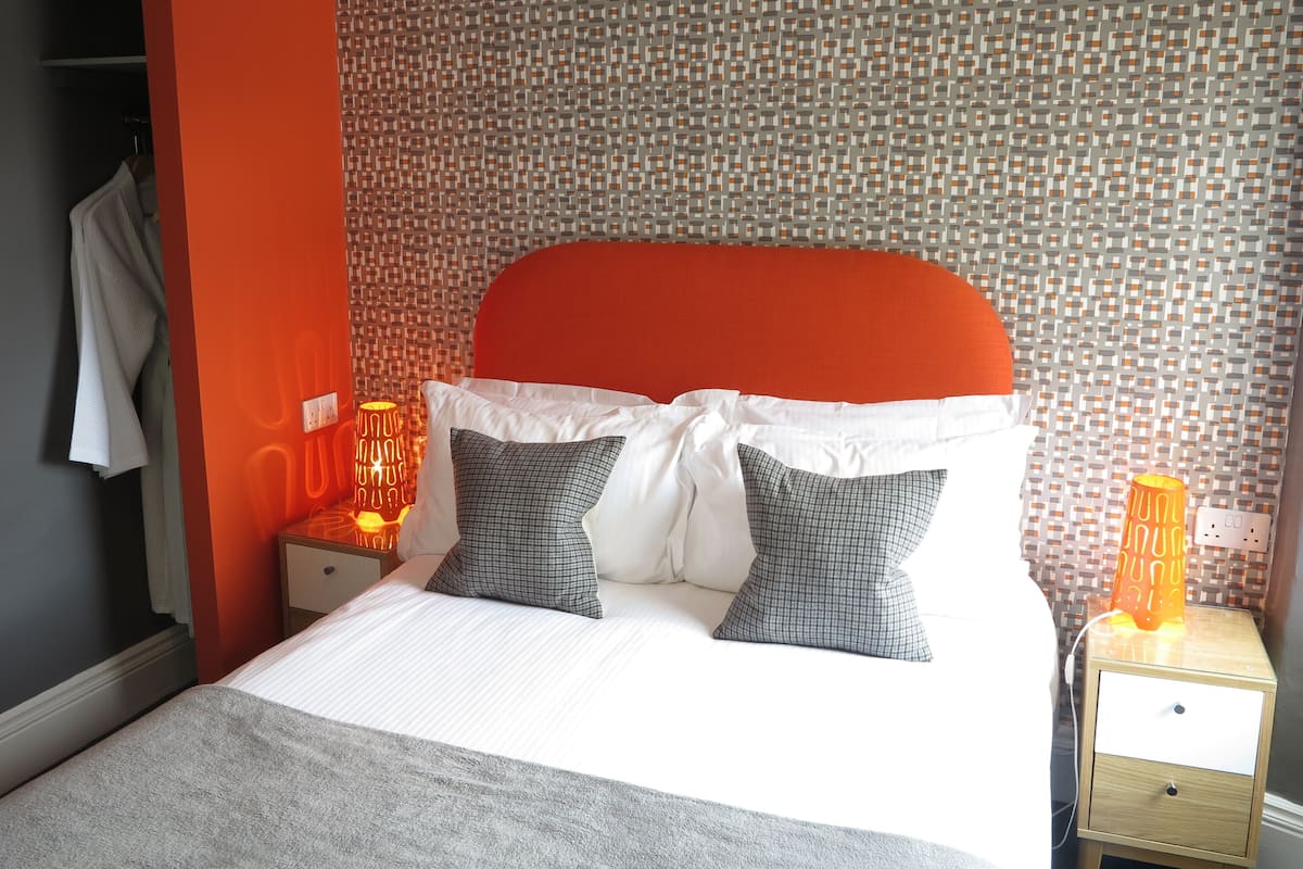 Un lit bien fait, avec une tête de lit rouge, des oreillers blancs et gris, et deux lampes de chevet.