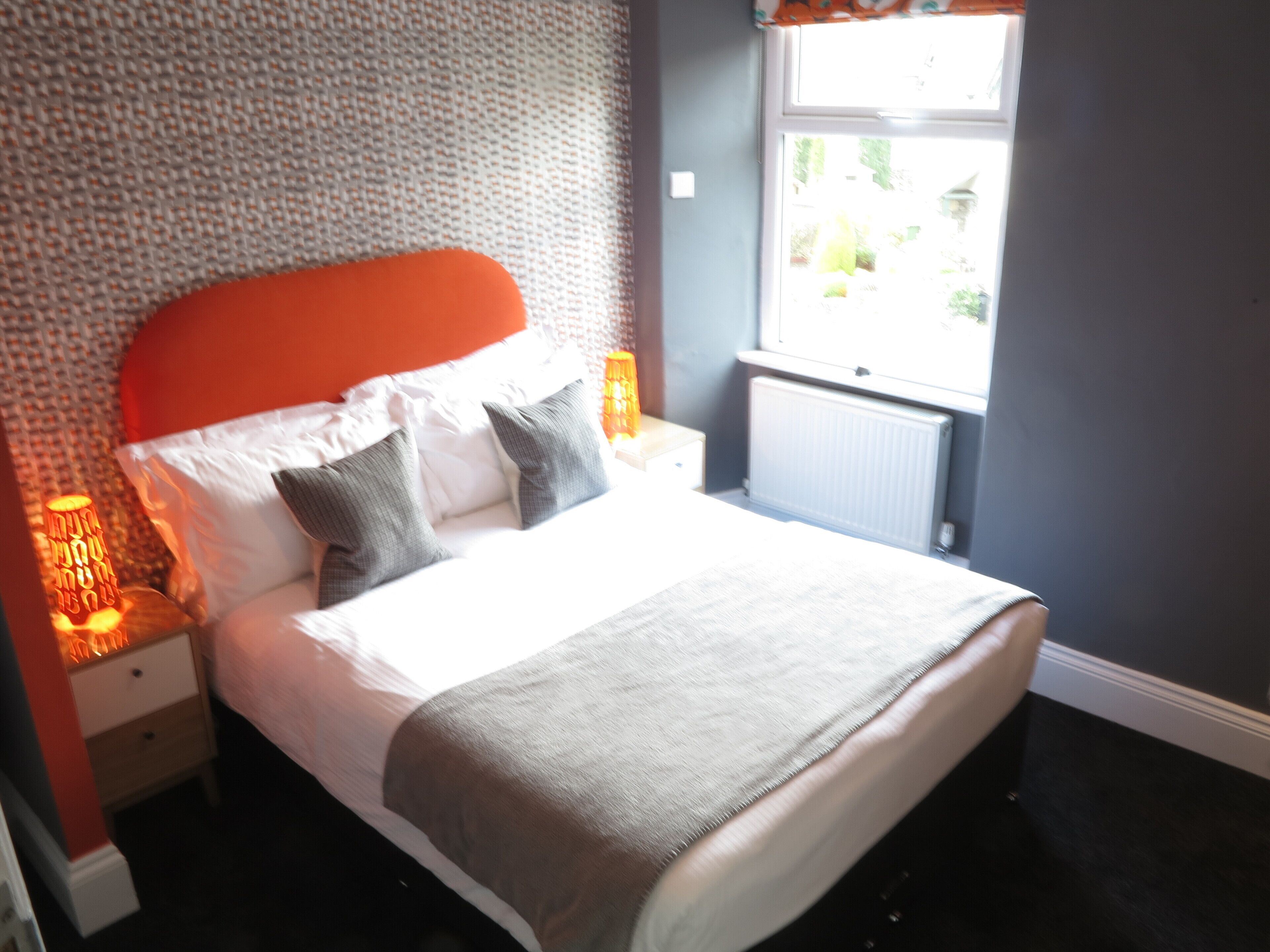 A neatly made bed with pillows and a blanket, a nightstand with a lamp, and a window with a view of greenery outside.