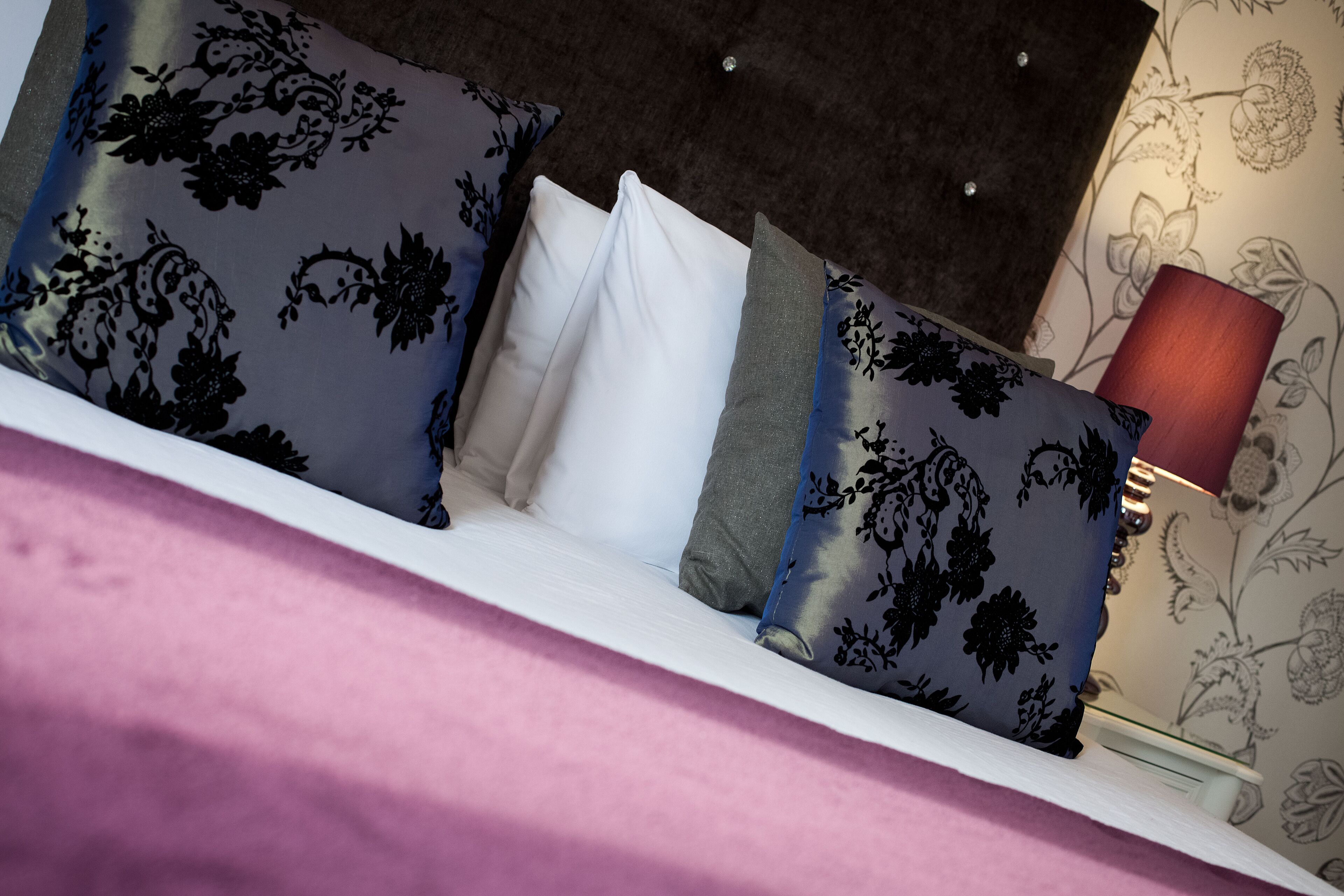 Un lit avec des oreillers à motifs floraux, un oreiller blanc et une lampe rouge, le tout placé près d’un mur recouvert d’un papier peint à fleurs.